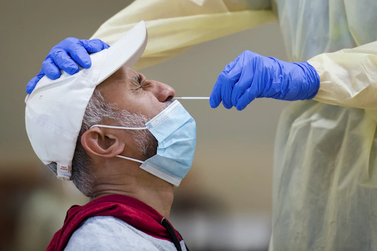 A man in profile wearing a white baseball cap and a face mask pulled down below his nose, winging as the gloved hand of a health-care worker inserts a test swab into his nose.