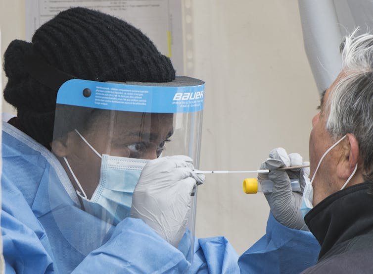 A female health-care worker in PPE holds a test vial and deploys a tongue depressor in one hand while holding a swab near his face with the other.