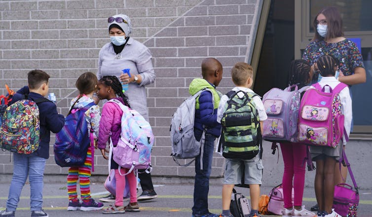 Teachers wearing face masks greet students in a school yard.