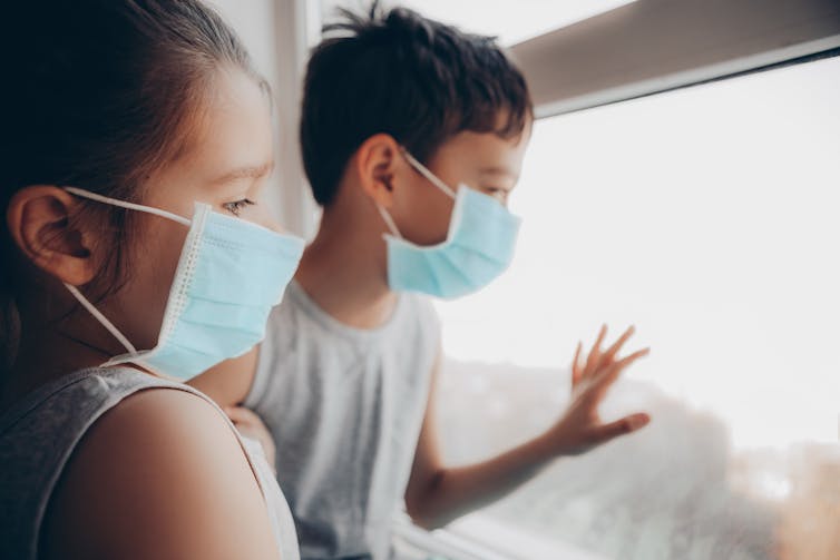 Two children wearing masks looking out a window
