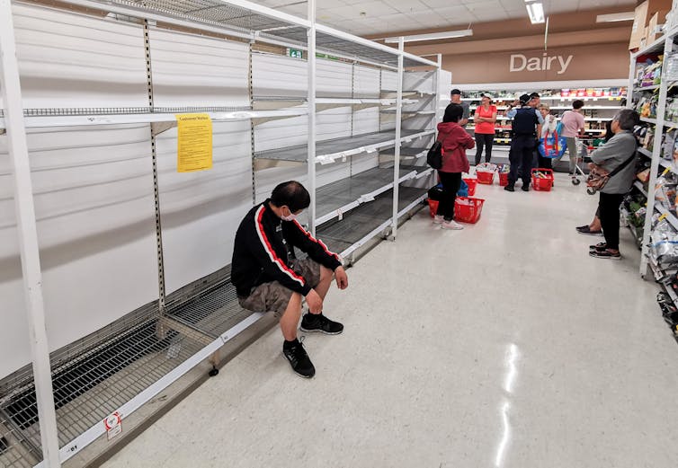 A man sits on an empty supermarket shelf.