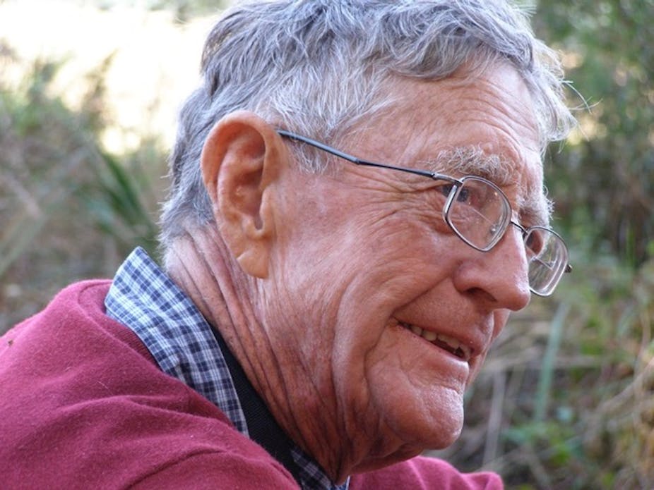 Profile of a man with a half-smile, spectacles, grey hair and with a maroon jersey on.