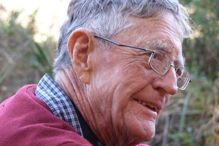 Profile of a man with a half-smile, spectacles, grey hair and with a maroon jersey on.