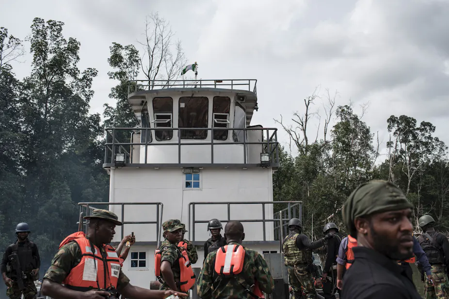 tIn the foreground, a dozen men in uniform who stand on a boat with dense trees in the background.