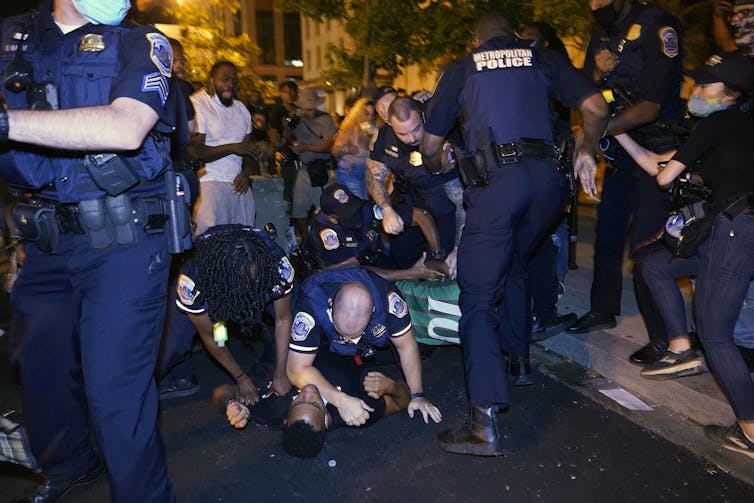Two police officers pin a Black man down on the ground, surrounded by other police officers and protestors.