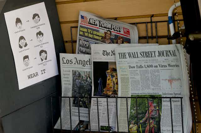 Newspapers for sale on a rack next to a sign exhorting people to wear a mask