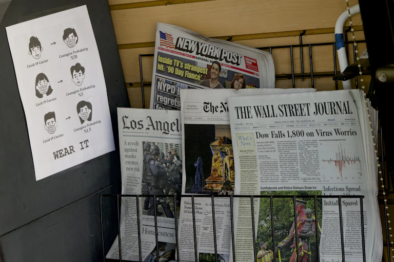 Newspapers for sale on a rack next to a sign exhorting people to wear a mask
