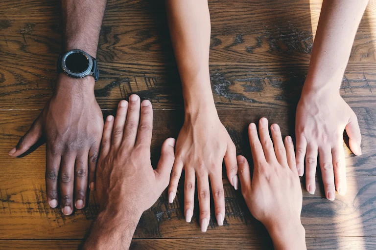 Five hands of different hues on a wooden table.