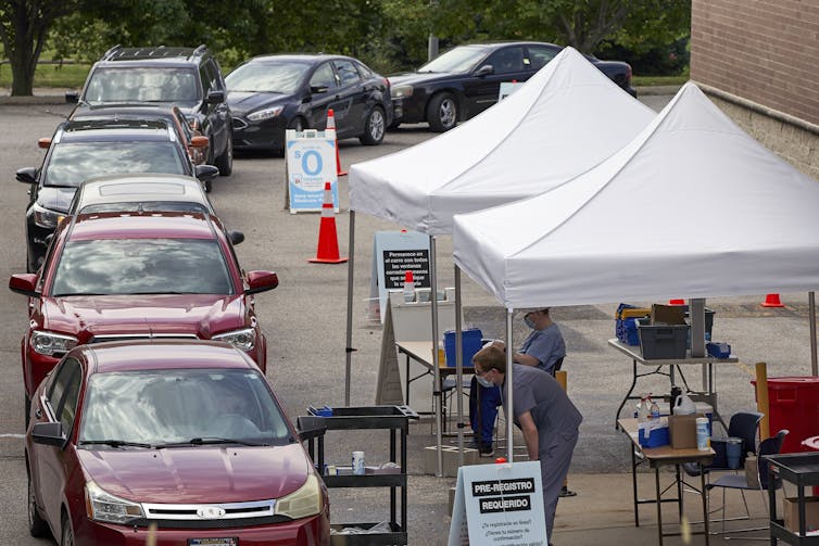 People waiting in a line of cars with health care workers standing under pop-up shade structures.