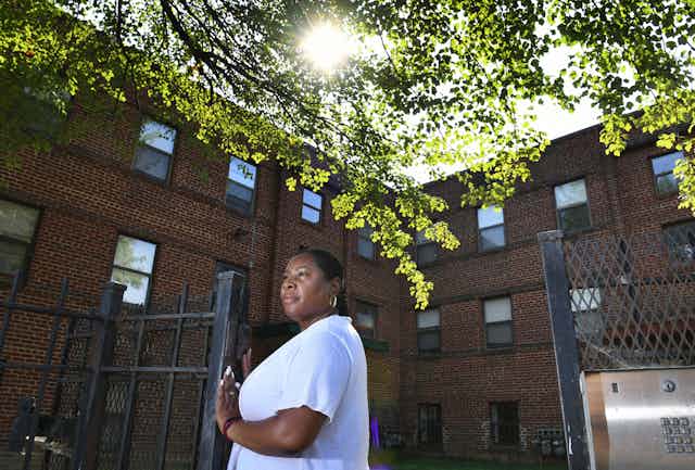 A Black woman stands at a gate to an apartment building.