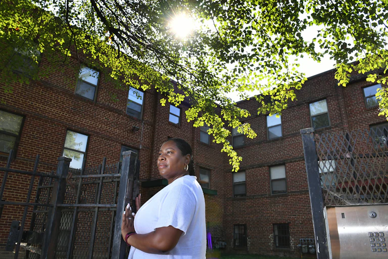 A Black woman stands at a gate to an apartment building.