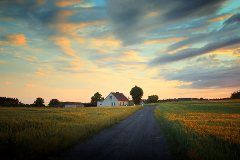 A country road leading past a white farmhouse with a red roof.