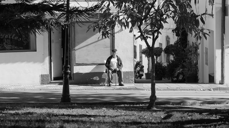 Foto en blanco y negro de un hombre mayor sentado en una silla, en la acera de un barrio de casas blancas.