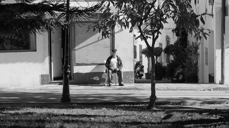 Foto en blanco y negro de un hombre mayor sentado en una silla, en la acera de un barrio de casas blancas.