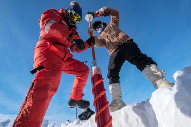 two scientists fully covered in cold weather clothing drill into the ice to extract an ice core. The photo is taken looking up at the two scientists and the photographer appears to be inside a pit dug into the ice.