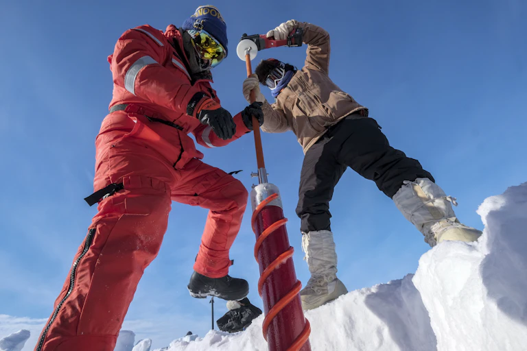 two scientists fully covered in cold weather clothing drill into the ice to extract an ice core. The photo is taken looking up at the two scientists and the photographer appears to be inside a pit dug into the ice.