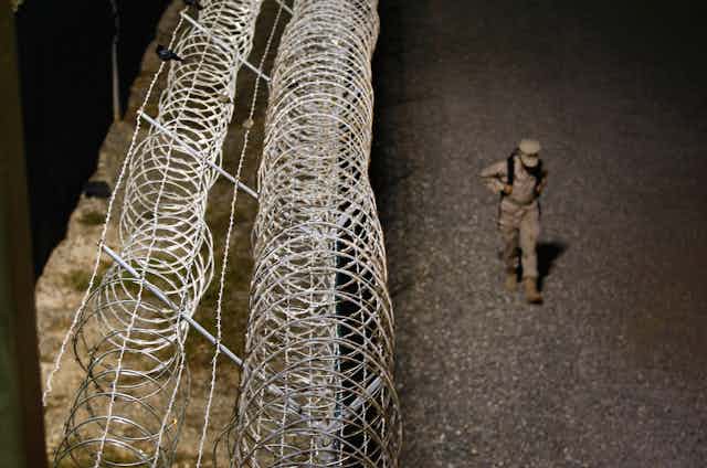 A uniformed soldier wearing a backpack walks next to the barbed wire fence surrounding Guantanamo Bay military base at night