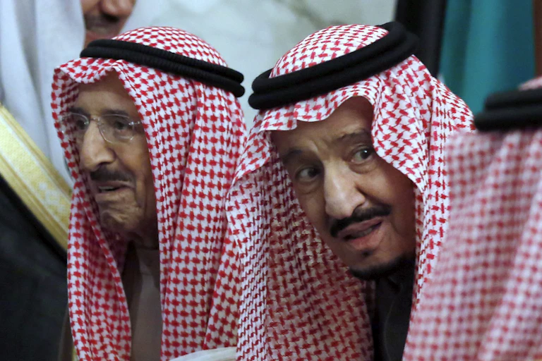Two men wearing head scarves sit together in mid-conversation..