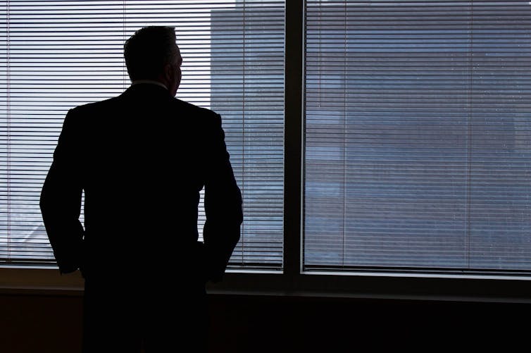 A man in silhouette in a suit stares out an office window.