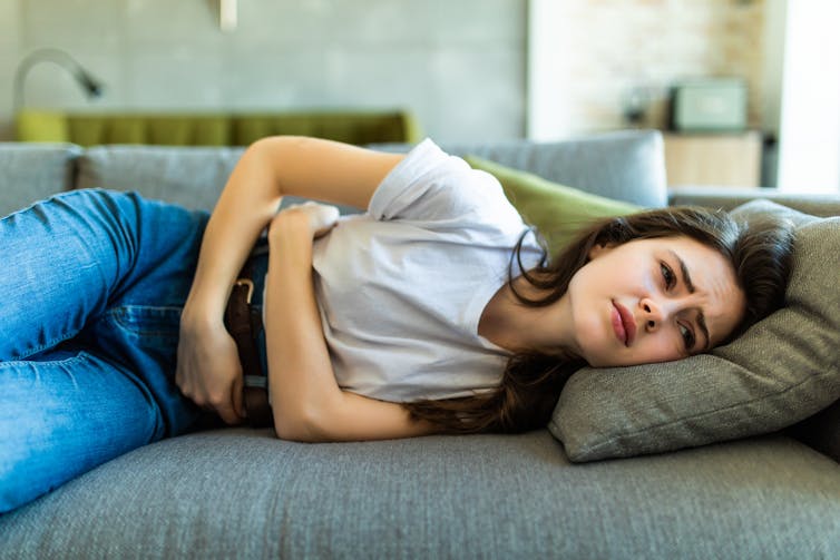 University of Sydney - A young woman lies on the couch clutching her stomach. Her facial expression indicates discomfort.