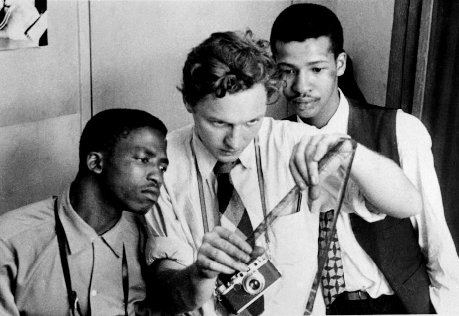 Three men in 1950s fashions examine a roll of film emerging from a camera.