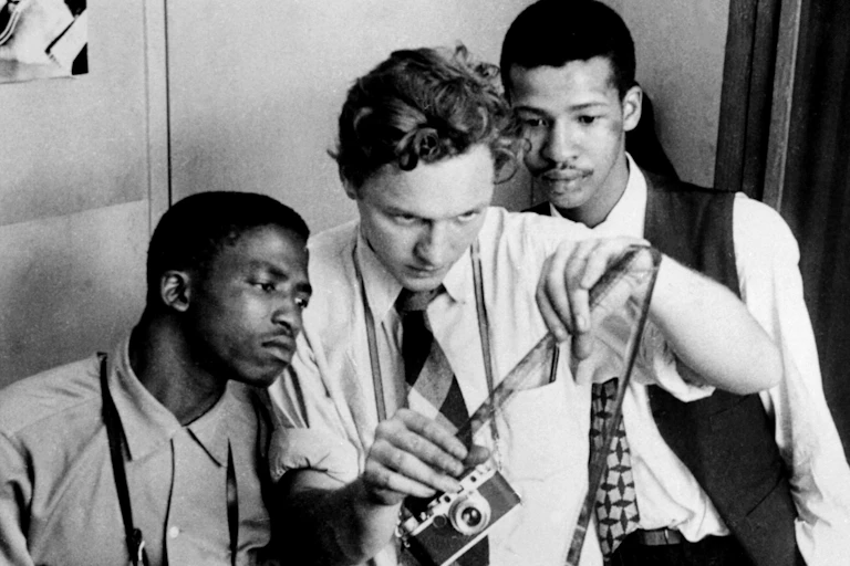 Three men in 1950s fashions examine a roll of film emerging from a camera.