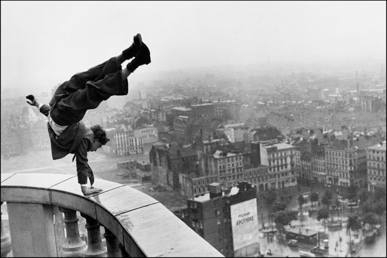 A man in a suit doing a one-armed handstand on a rain-wet balcony railing high above a city, his legs curling over towards the city.