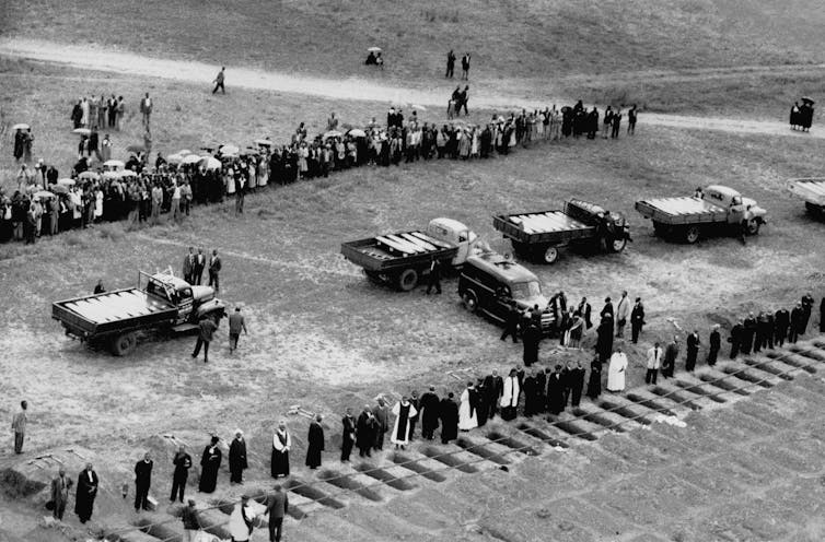 Aerial view of five trucks carrying rows of coffins in the centre of a field. Mourners and clergy stand at a distance on either side, alongside dozens of graves, some still empty.