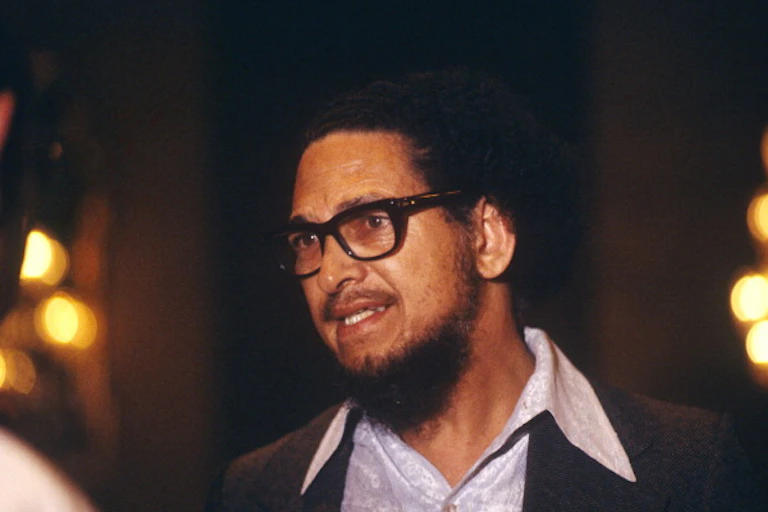 A bearded man with dreadlocks and heavy-framed spectacles gazes intently, looking ready to speak.