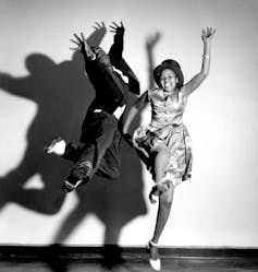 A smartly dressed male and female couple dance, hands and feet in the air, the male dancer mid-air, with their shadows against a white wall.