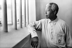 A man with greying hair leans his arm against a window sill as he gazes out through bars on the window.