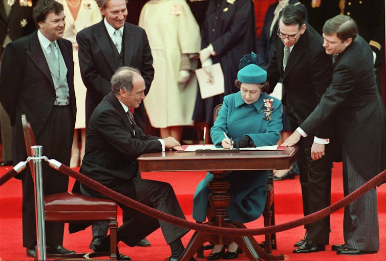 Pierre Trudeau sits at a desk on a red carpet looking on as Queen Elizabeth, in a teal suit, signs a proclamation.