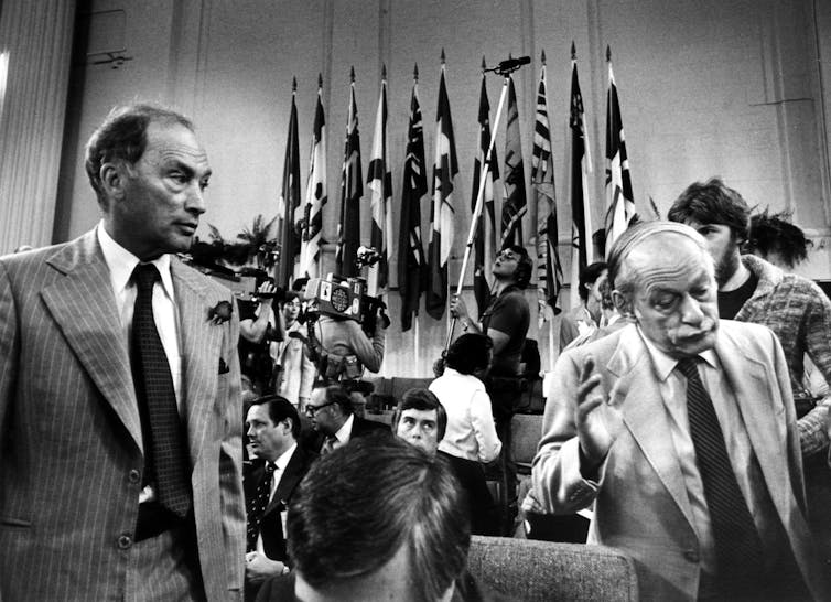 Rene Levesque, Québec premier, shrugs his shoulders and walks away from Pierre Trudeau at a conference with flags in the background.