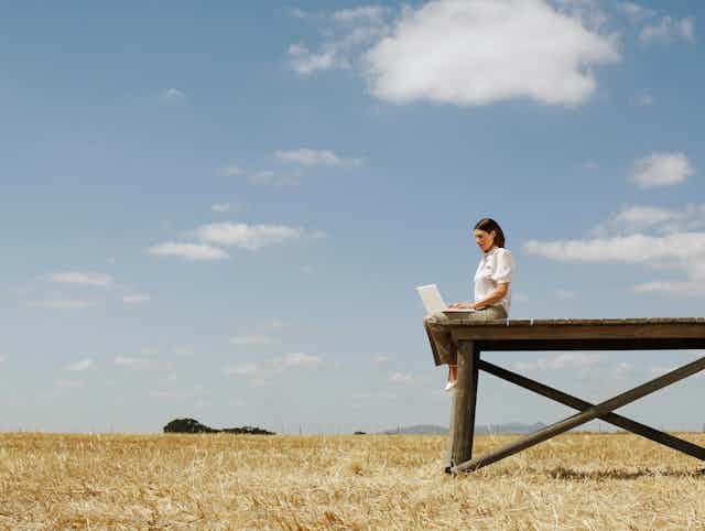 A woman with a laptop on her lap sits on a wooden structure in an open field.