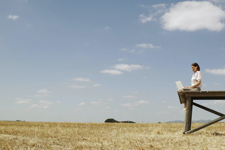 A woman with a laptop on her lap sits on a wooden structure in an open field.