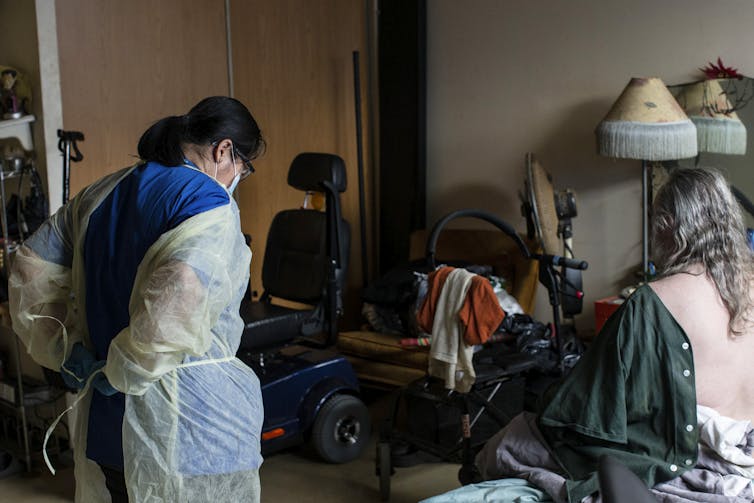 A personal support worker puts on a plastic robe while her client sits on the bed waiting for her help. Wheelchairs sit next to the bed.