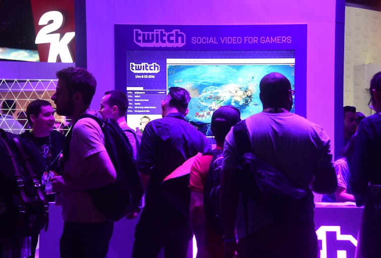 A group of youths stand under purple lights in front of a Twitch booth at a video game convention.