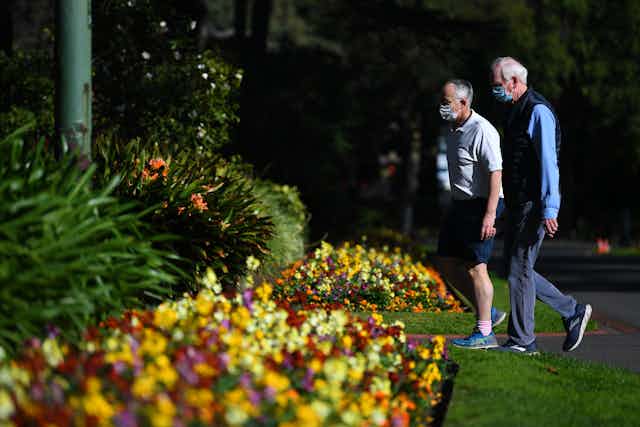 Two men wearing masks walk through Fitzroy Gardens in Melbourne on a sunny day.