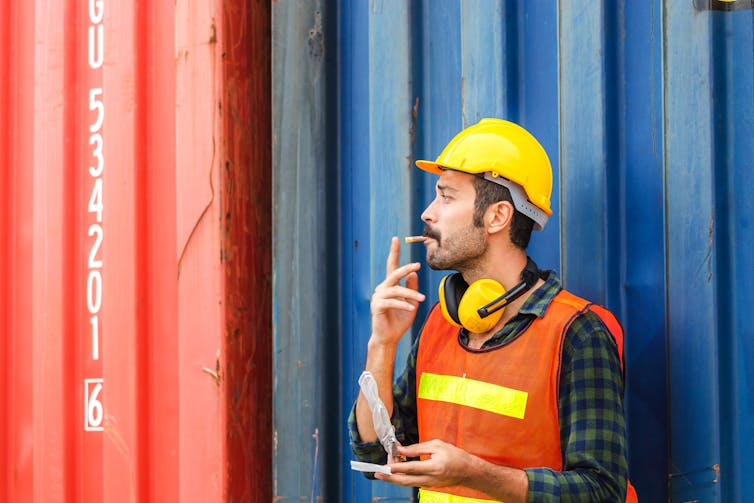 Worker in high-viz jacket and hard hat smoking as he leans against a shipping container