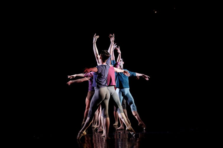 Ballet dancers seen from behind, in a row with toes pointed and arms outstretched.