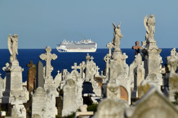 A cruise ship is seen at sea, framed by gravestones on land.