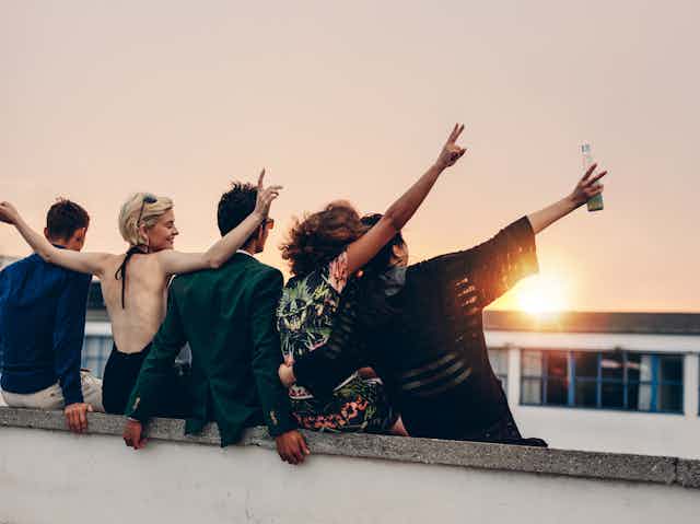 Five young people, with backs to camera, hugging, arms in the air, looking at the sunset
