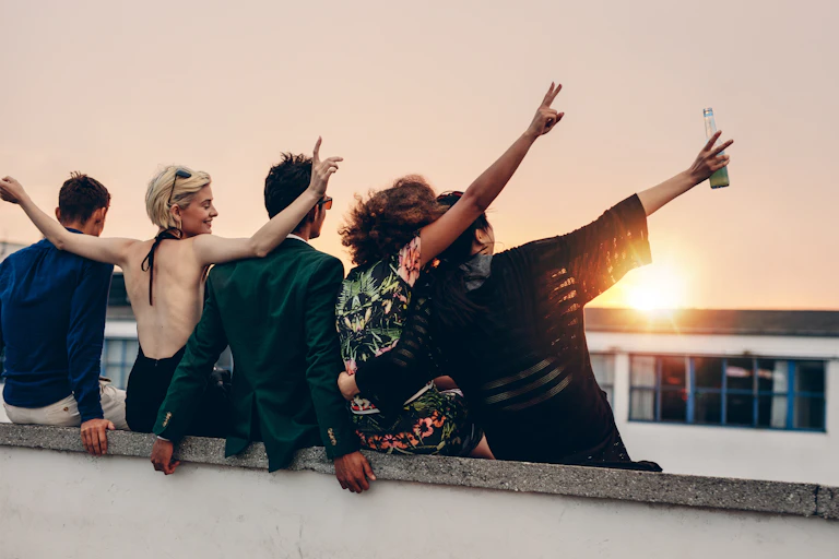 Five young people, with backs to camera, hugging, arms in the air, looking at the sunset