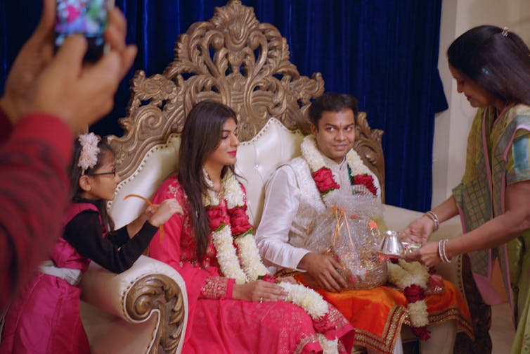 A couple sitting on a throne being presented with gifts.