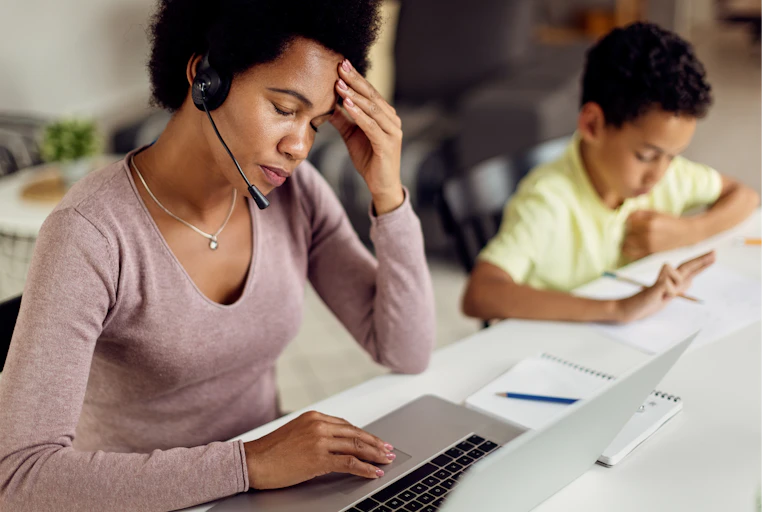 A Black woman working at a laptop and wearing a headset places a hand on her temple as her young son studies beside her.