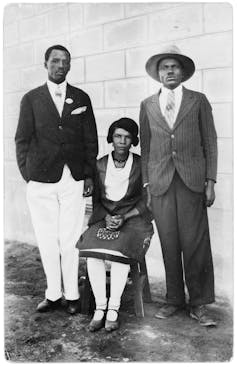 Two men stand and a woman sits between them on a chair, all in formal attire and looking at the camera unsmilingly.