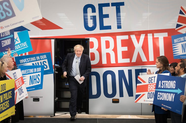 Boris Johnson steps off bus with words 'GET BREXIT DONE' surrounded by cheering crowd.