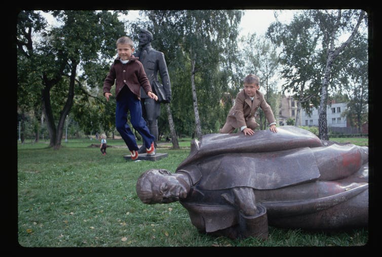 Two young blond boys jump and crawl on a fallen bronze statue in a park