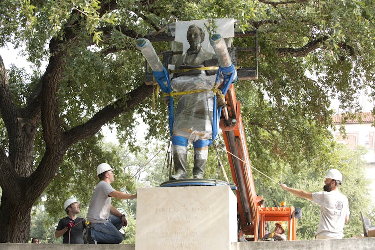 Workers in hardhats use a small orange crane to hoist a plastic-wrapped and padded bronze statue