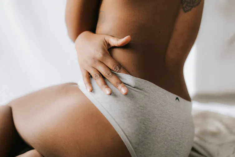 The back of a woman, one hand touches the top of her underwear. She wears nothing else.